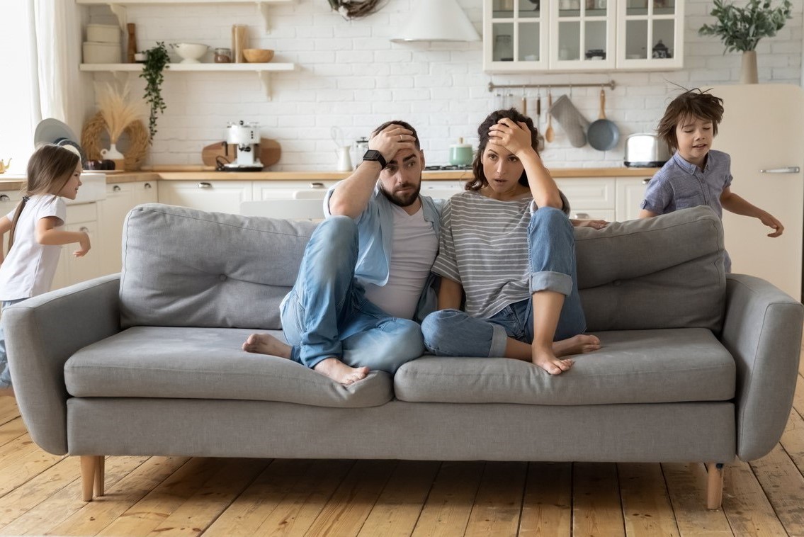 Tired white mother and white father sitting on couch with hands on heads while daughter and son run around sofa where parents are sitting.
