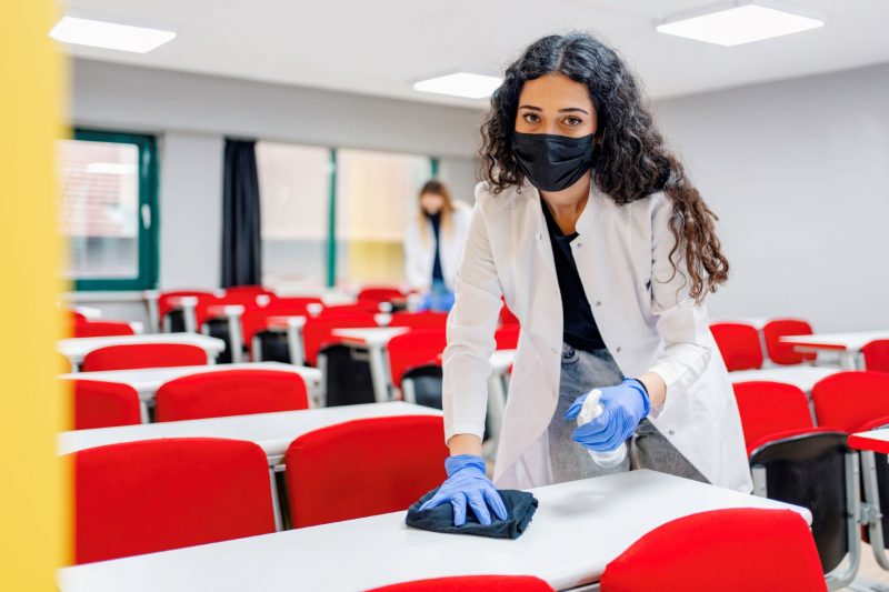 A teacher cleaning a desk in a classroom while wearing a face mask