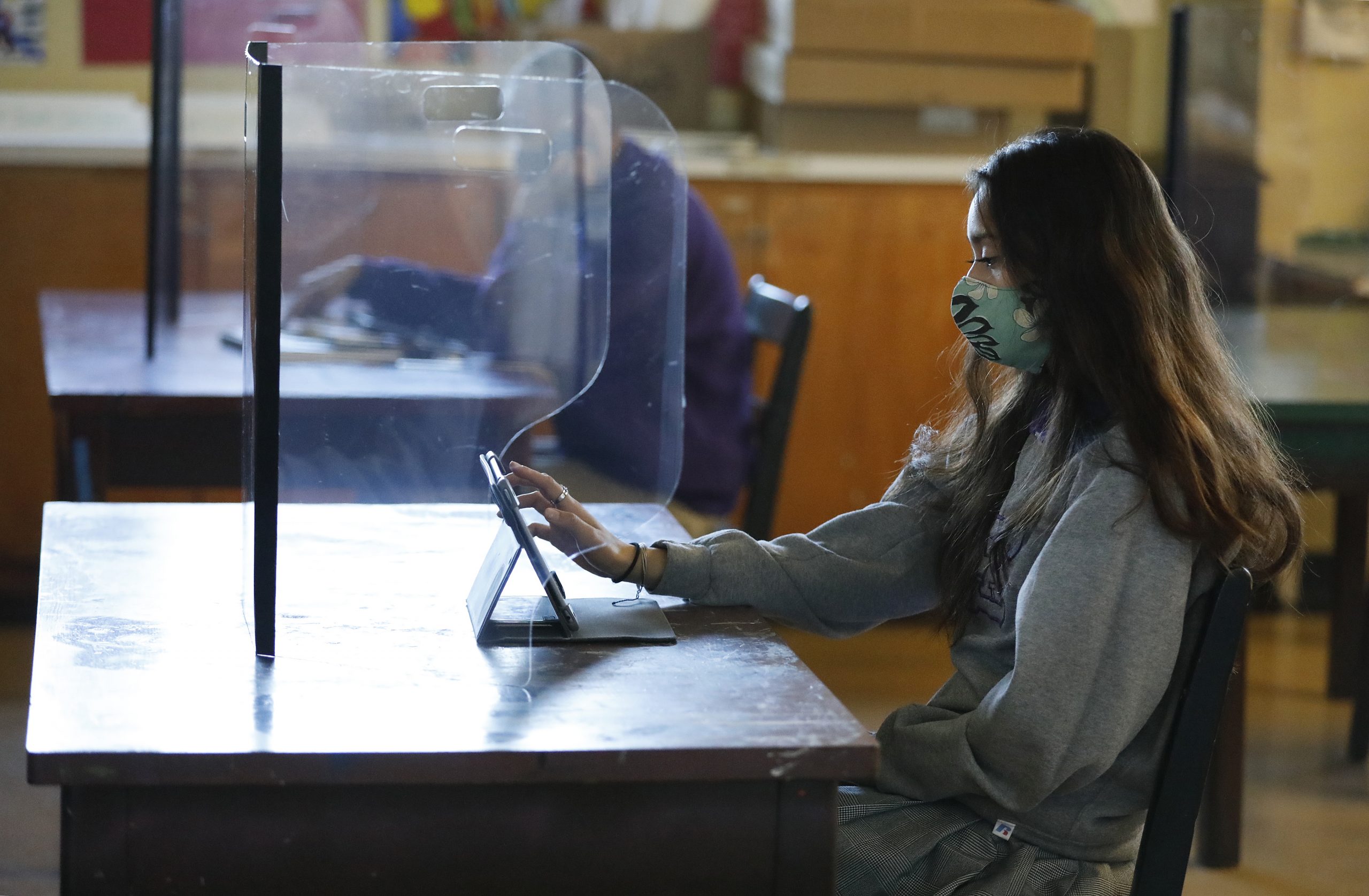 Side view of older teen girl with long brown hair, wearing mask sits at desk with a plexiglass barrier in classroom. She is using a tablet.