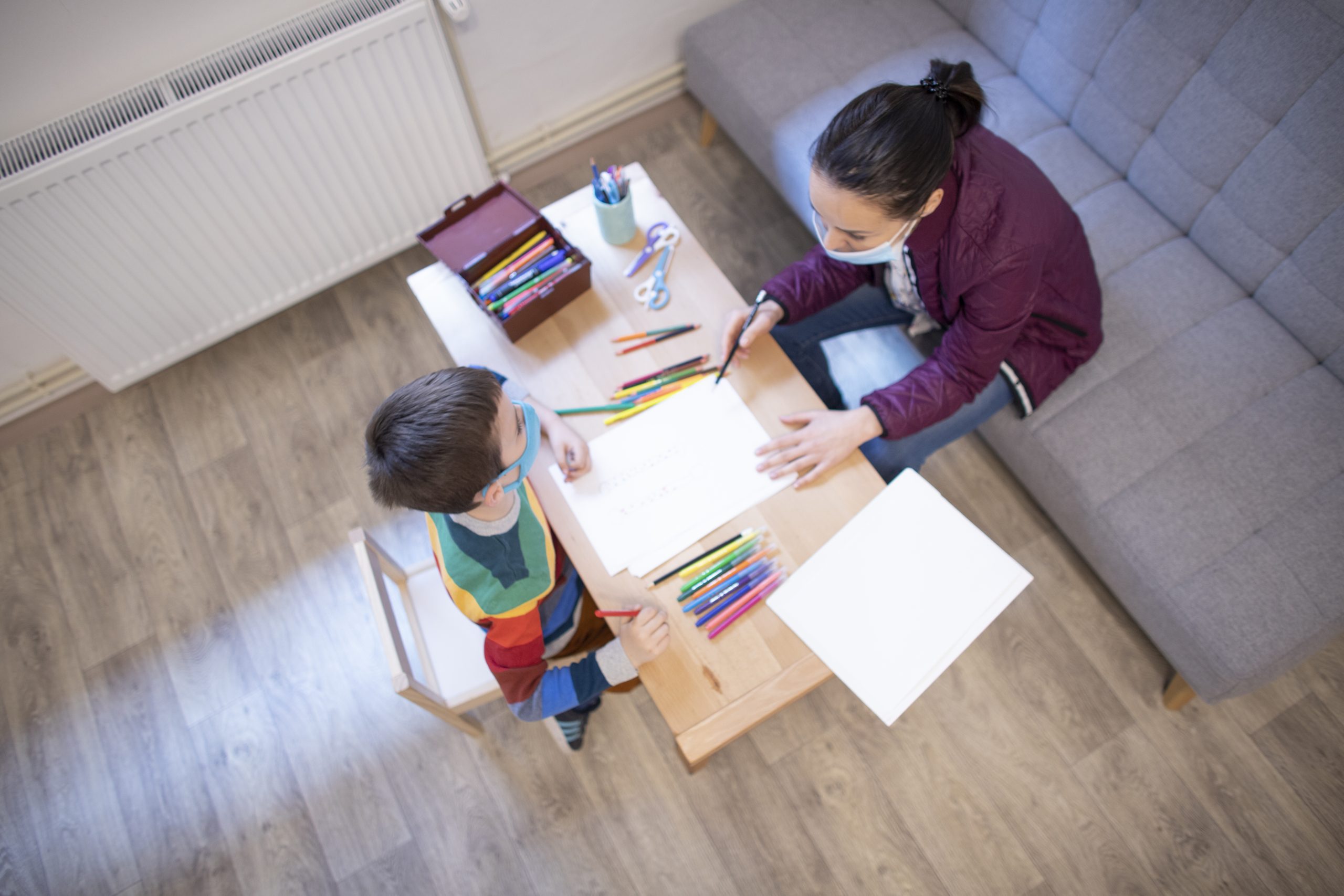 Bird's eye view of six year old white boy wearing mask working with a white, female psychologist wearing a mask at the psychotherapy session..