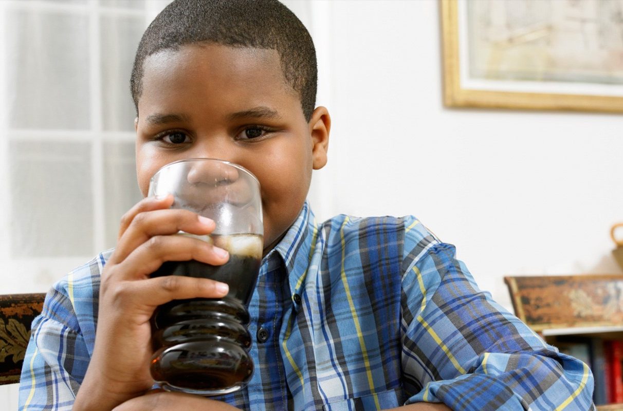 Black boy drinking soda from a glass. He wears a blue plaid shirt and is smiling.
