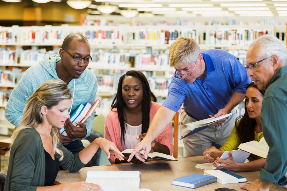 a multi-ethnic group of adults meeting in a library. Mixed ages, 20s to 70s. They are all sitting or standing around a table holding books. Everyone is looking at the book that a man in a blue shirt is pointing to.