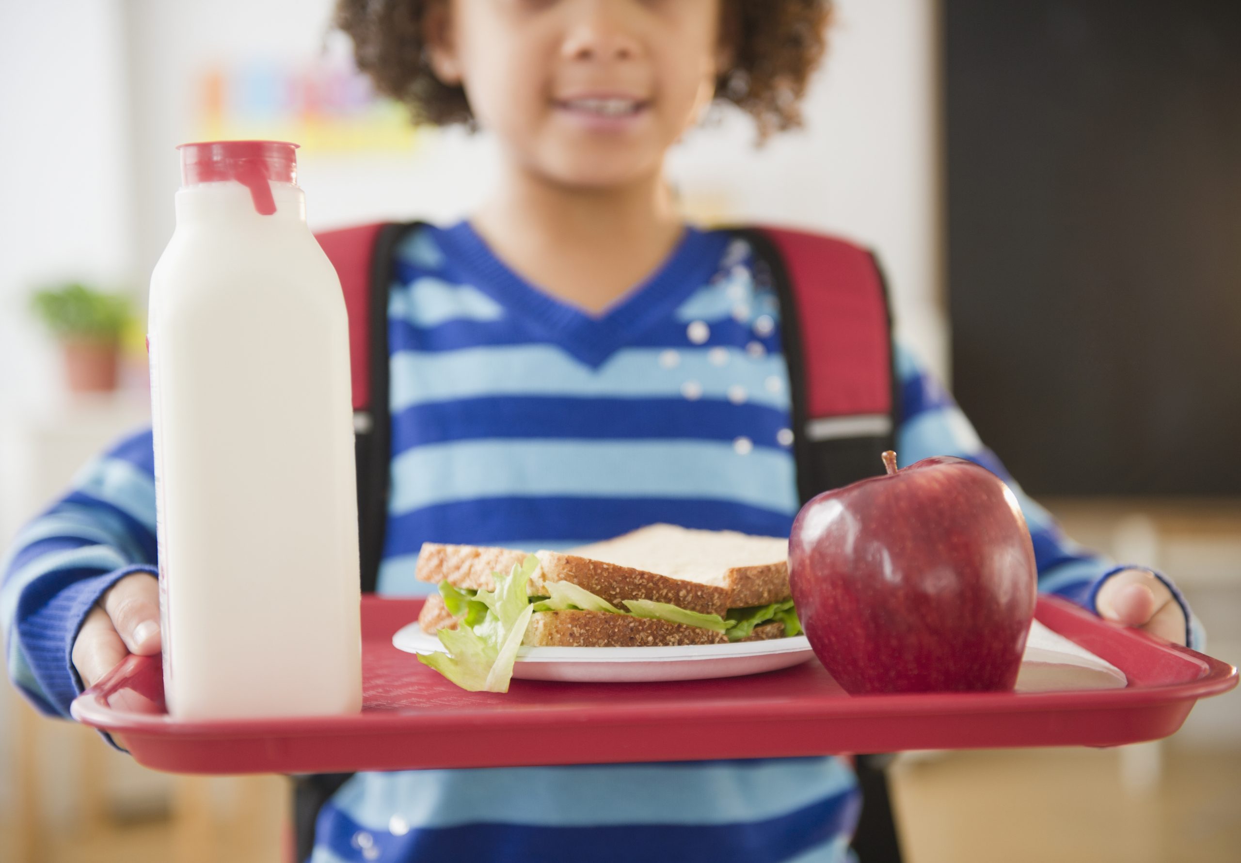 A girl with a backpack on her back holds a tray in front of her with a sandwich, apple and bottle of milk