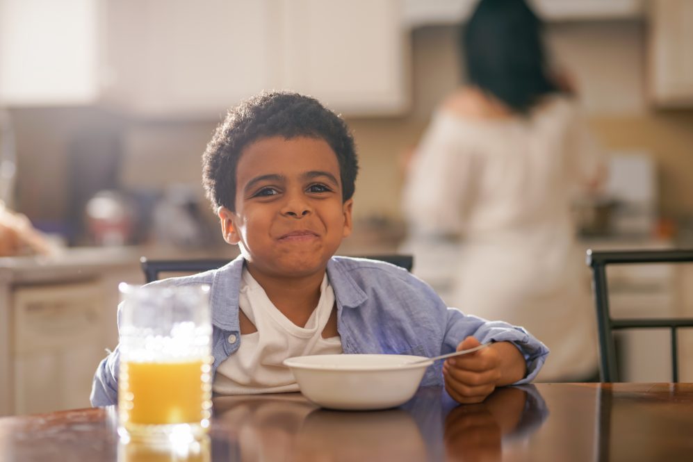 young boy sits at table eating cereal. He is looking at the camera and smiling. There is a glass of orange juice on the table.
