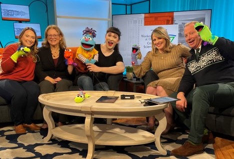 five adults sit on couches holding puppets and smiling