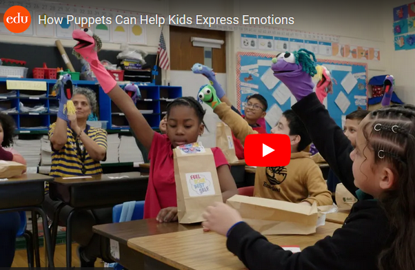 Screenshot of Video of diverse kids and teacher in classroom holding sock puppets up with one hand