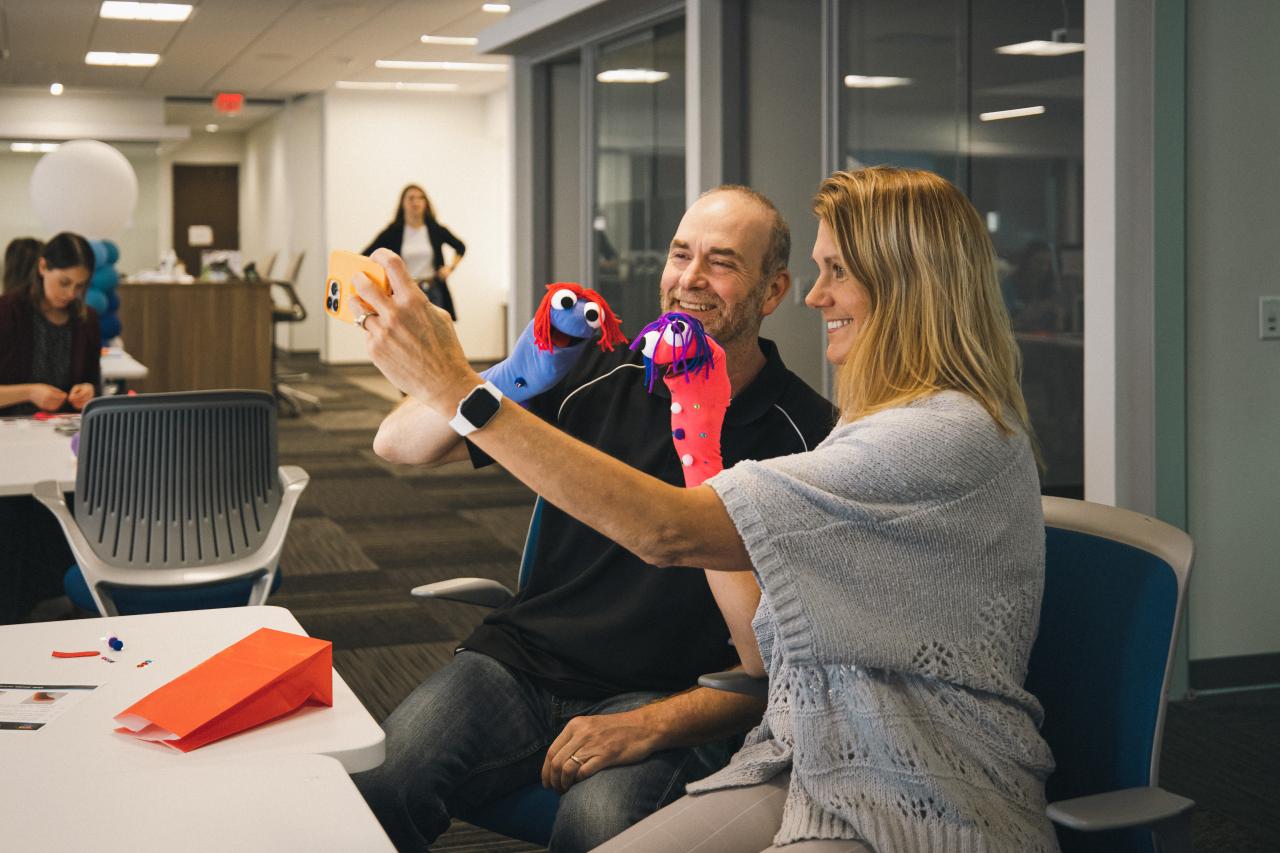A man and woman sit at a table in a workspace, taking a selfie with two sock puppets and smiling