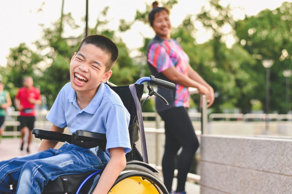 child on wheelchair wearing light blue polo shirt and jeans is laughing in outdoor park 